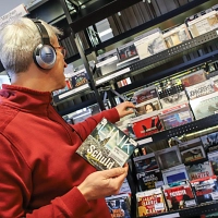 Ein Mann steht in der Stadtbücherei vor einem Regal mit Hörbüchern und trägt Kopfhörer. Er hält eine CD in der Hand.