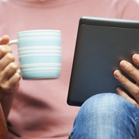 Eine Person sitzt in einem Sessel, in der einen Hand einen E-Book-Reader, in der anderen Hand eine Tasse