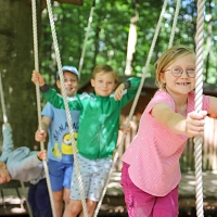 Ein Mädchen und drei Jungen halten sich an Tauen Seilen fest, sie balancieren auf hängenden Holzbalken im Stadtwald. Im Hintergrund sind Bäume zu sehen.