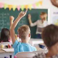 Kinder, die sich melden, sitzen in einem Klassenraum auf ihren Stühlen. Der Blick geht über ihre Köpfe hinweg nach vorne zur Tafel, wo die Lehrerin steht.