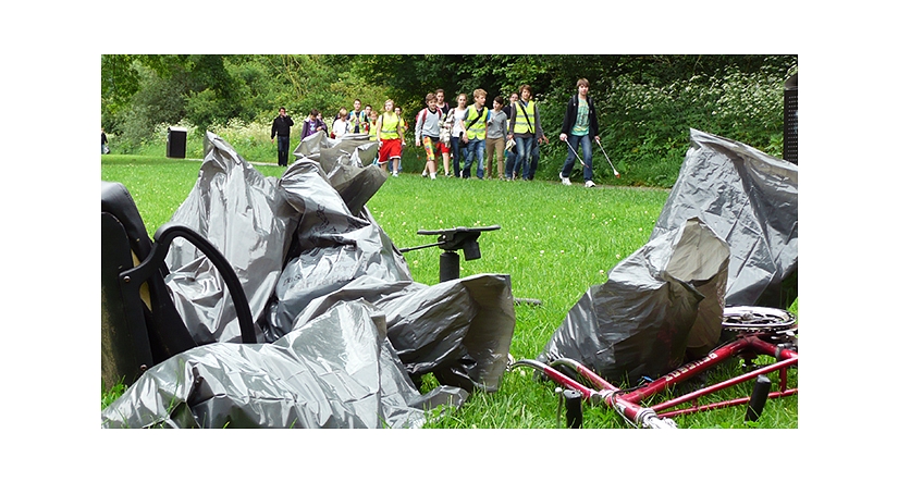 Das Foto zeigt Abfallsäcke und einen Fahrradrahmen ohne Reifen im Vordergrund. Dahinter gehen Kinder in Warnwesten mit Papierzangen und sammeln Abfälle ein. © DBM, Sonja Stender